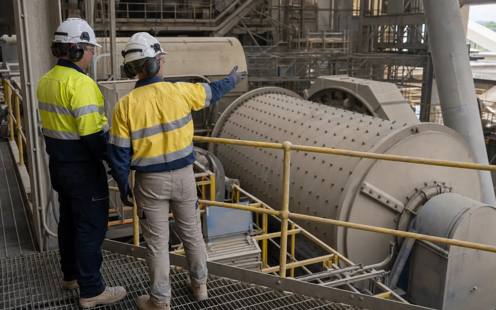 Two industrial professionals in full PPE safely reviewing a horizontal ball mill from a guarded access platform.